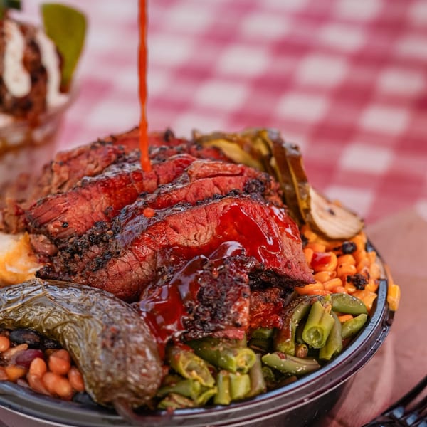 Brisket BBQ bowl with sliced meat, beans, corn, green beans, and peppers, drizzled with sauce.