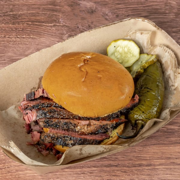 Brisket BBQ sandwich with sliced pickles and a roasted green pepper in a paper tray.
