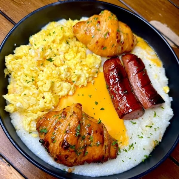 A black bowl filled with grits, scrambled eggs, two sausages, and two glazed croissants.