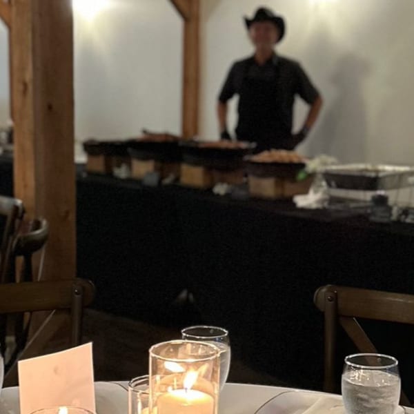 A server in a cowboy hat stands behind a buffet table with chafing dishes, with a candlelit table in the foreground.