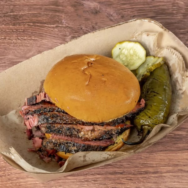 Brisket BBQ sandwich with pickles and a roasted green pepper in a paper tray on a wooden table.