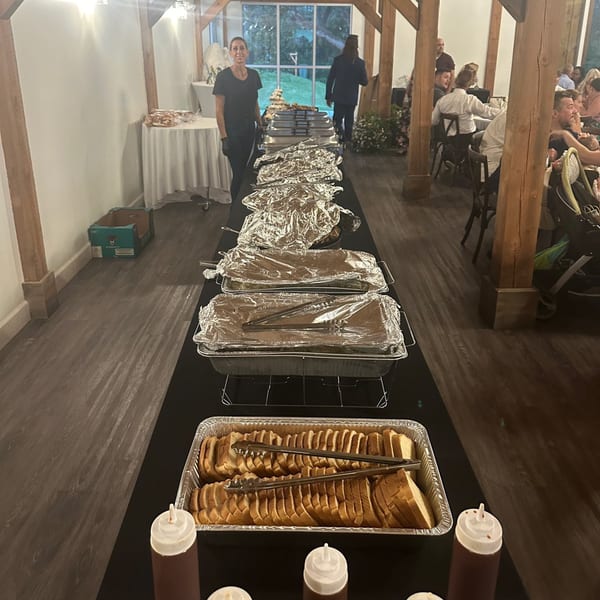 A buffet line with foil-covered trays, sliced bread, and a server in a rustic event space.